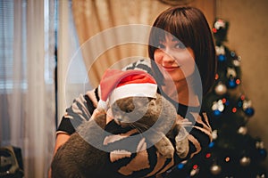 Woman with her cat wearing Santa Claus hat near christmas tree