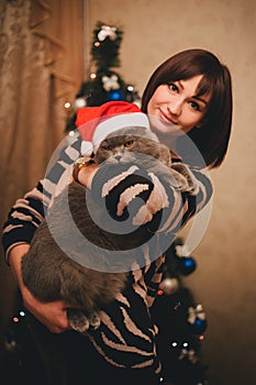Woman with her cat wearing Santa Claus hat near christmas tree