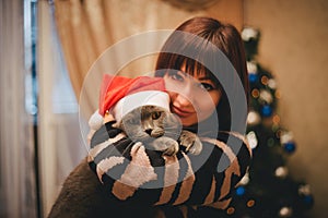 Woman with her cat wearing Santa Claus hat near christmas tree