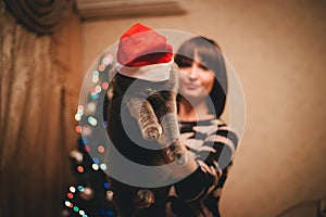 Woman with her cat wearing Santa Claus hat near christmas tree
