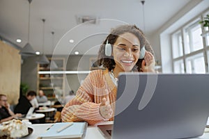 A woman at her desk in front of a laptop computer with headphones, smiling in a cozy minimalistic cafe