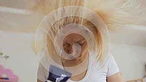 A woman in her forties dries her dyed blond hair with a hair dryer.