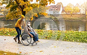 Woman and her friend with face mask in a wheelchair having stroll through the park at a river enjoying the autumn during covid-19