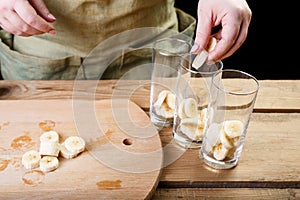 The woman with her hand puts the pieces of bananas into three empty glass glasses