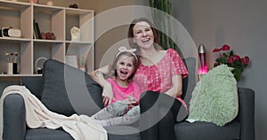 Woman and her little daughter sitting on the sofa and watching comedy film on TV laughing.