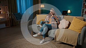 A woman and her young daughter are watching TV using a remote control. Mom and daughter sitting in an embrace in the