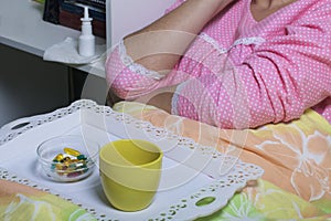 A woman hesitates before taking medication. He lies in bed under the blanket. Nearby is a tray with pills and a glass of water.