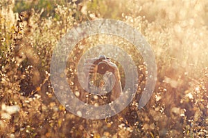 Woman hidden in tall grass field holding camera in hands and taking pictures