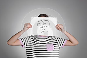 Woman hiding behind sheet of paper with sad face on grey background