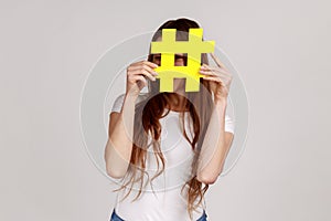 Woman hiding behind yellow hashtag symbol, covering face with hash sign, viral topics on internet.