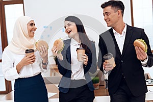 A woman in a hijab with her colleagues eating a hamburger and drinking coffee.
