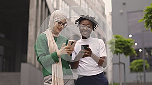 Woman in hijab withsmartphone and african female with phone standing together near office building