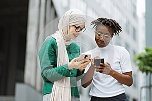 Woman in hijab withsmartphone and african female with phone standing together near office building