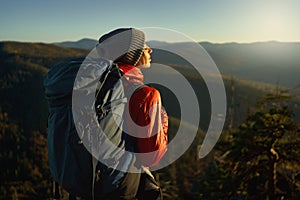 Woman hiker with backpack standing on edge of cliff against sunset sky over mountains