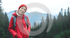 Woman hiker with backpack, wearing in red jacket and cap, standing on the forest and mountains background