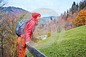 Woman hiker with backpack, wearing in red jacket and orange pants, standing on the mountains background