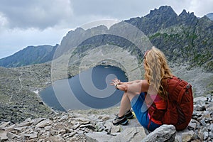 Woman hiker with curly blonde hair and red backpack looks towards Vysne Wahlenbergovo lake in Slovakia High Tatras mountains.