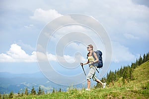 Woman hiker hiking on grassy hill, wearing backpack, using trekking sticks in the mountains