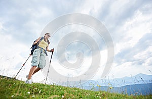 Woman hiker hiking on grassy hill, wearing backpack, using trekking sticks in the mountains