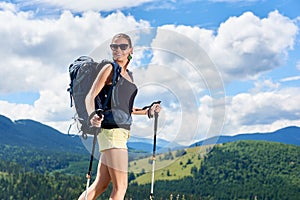 Woman hiker hiking on grassy hill, wearing backpack, using trekking sticks in the mountains