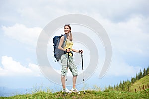 Woman hiker hiking on grassy hill, wearing backpack, using trekking sticks in the mountains