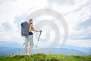 Woman hiker hiking on grassy hill, wearing backpack, using trekking sticks in the mountains