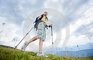 Woman hiker hiking on grassy hill, wearing backpack, using trekking sticks in the mountains