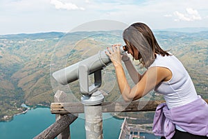 Woman hiker at mountain viewpoint looking throgh touristic telescope at the lake and canyon in the distance