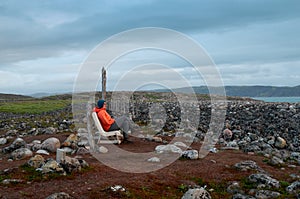 A woman hiker in an orange jacket sits on a plastic chair on a rocky ocean shore