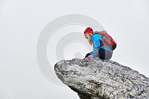 Woman hiker with red backpack sits at the edge of a cliff and looks down, isolated by fog. Hiking in Slovakia High Tatras