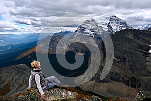 Woman hiker sitting on mountain top looking at  view of mountain valley with yellow larch trees from above.