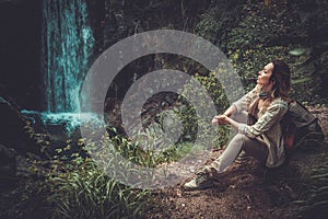 Woman hiker sitting near waterfall in deep forest.