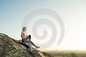 Woman hiker sitting on a steep big rock enjoying warm summer day. Young female climber resting during sports activity in nature.