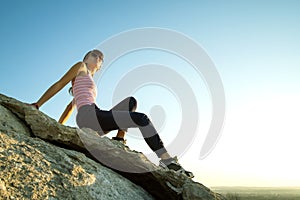 Woman hiker sitting on a steep big rock enjoying warm summer day. Young female climber resting during sports activity in nature.