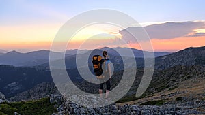 A woman hiker stands on a large rock with the mountains in the background and watches the sunset.