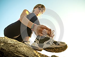 Woman hiker tying shoe laces of her sport boots while climbing steep big rock on a sunny day. Young female climber overcomes