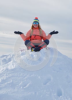 Woman hiker wearing warm Winter clothes sits on top of snow pile with her arms stretched out and her legs crossed, happy.