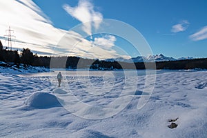 Woman with hiking backpack on field of deep crystal snow with scenic view of frozen alpine lake Forstsee, Techelsberg, Austria