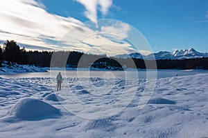 Woman with hiking backpack on field of deep crystal snow with scenic view of frozen alpine lake Forstsee, Techelsberg, Austria