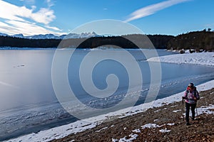 Woman with hiking backpack walking along shore of the frozen lake Forstsee, Techelsberg, Carinthia (Kaernten), Austria, Europe