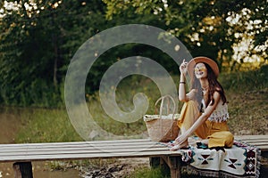 Woman in hippie eco-dress sits outdoors in the fall by the lake on a bridge resting, smiling and admiring the scenery