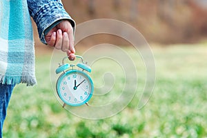 Woman holding alarm clock over spring flowers background. Daylight saving time reminder