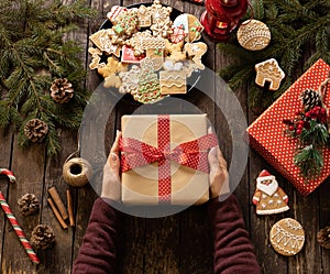 Woman holding a beautifully decorated Christmas gift on rustic wooden table next to a plate full of baked ginger cookies with