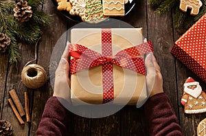 Woman holding a beautifully decorated Christmas gift on rustic wooden table next to a plate full of baked ginger cookies with