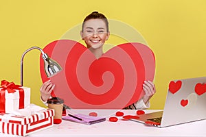 Woman holding big red paper heart, smiling, sitting at workplace with laptop in sticky hearts.