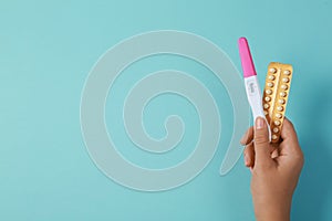 Woman holding birth control pills and pregnancy test on light blue, top view. Space for text