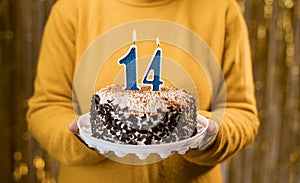 Woman holding birthday cake with burning candles number 14 against decorated background, close up. Celebrates birthday