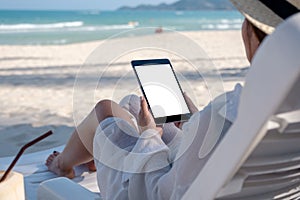 A woman holding a black tablet pc with blank desktop screen while laying down on beach chair on the beach