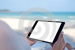 A woman holding a black tablet pc with blank desktop screen while laying down on beach chair on the beach