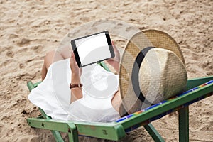 A woman holding a black tablet pc with blank desktop screen while lying down on a beach chair